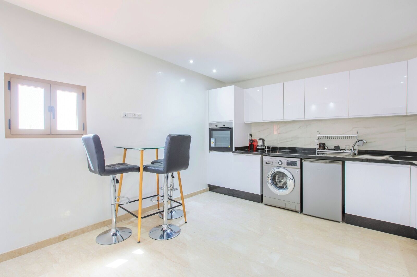 Una cocina moderna con armarios blancos, lavadora, microondas y un comedor con dos sillas y una mesa de vidrio.