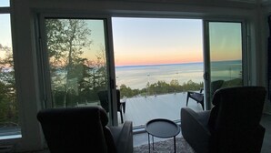 Une pièce avec deux chaises et une petite table, de grandes fenêtres donnant sur un plan d’eau et des arbres, et une porte en verre.