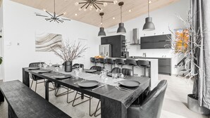 Une cuisine moderne dotée d’un îlot central, de luminaires suspendus et d’un coin repas avec une longue table pouvant accueillir six personnes.