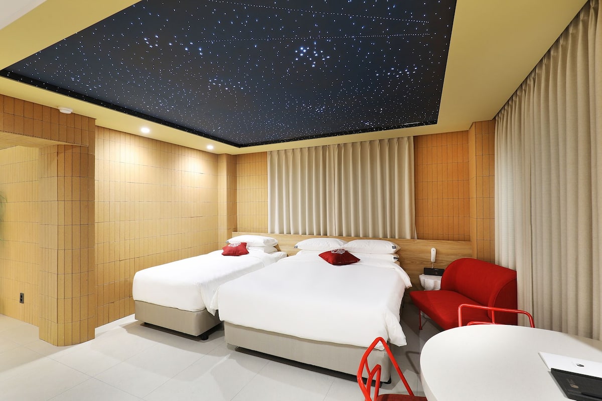 Ein Hotelzimmer mit Sternenhimmel, zwei Betten, einem roten Sessel und einem Schreibtisch.
