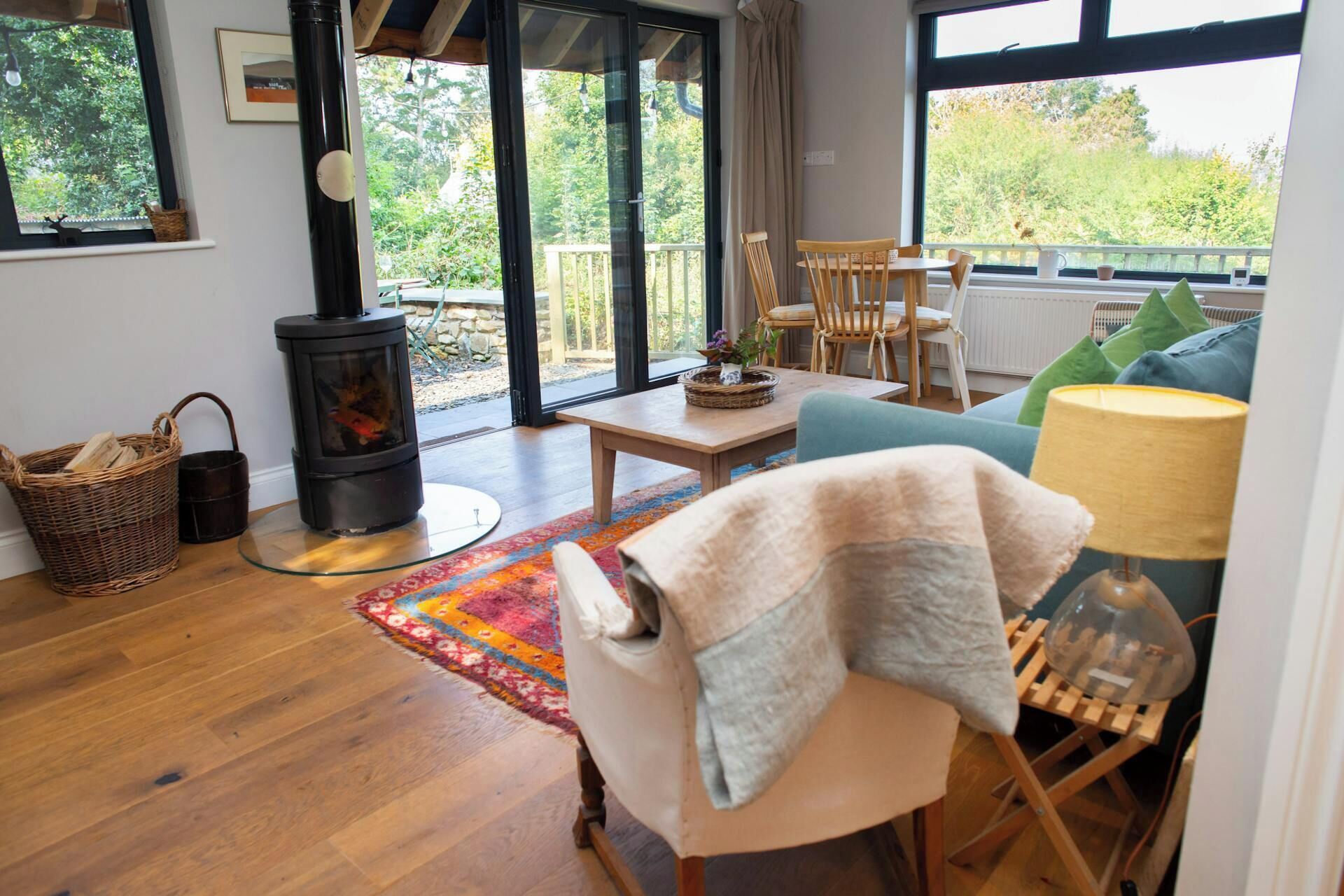 A cozy living room with a wood-burning stove, a sofa, a table, and a view of the outdoors through large windows.