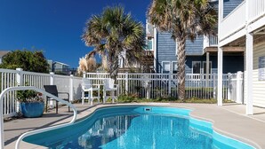 Ein Swimmingpool mit weißer Liegefläche, umgeben von einem weißen Zaun, Palmen und einem Gebäude mit Balkon.