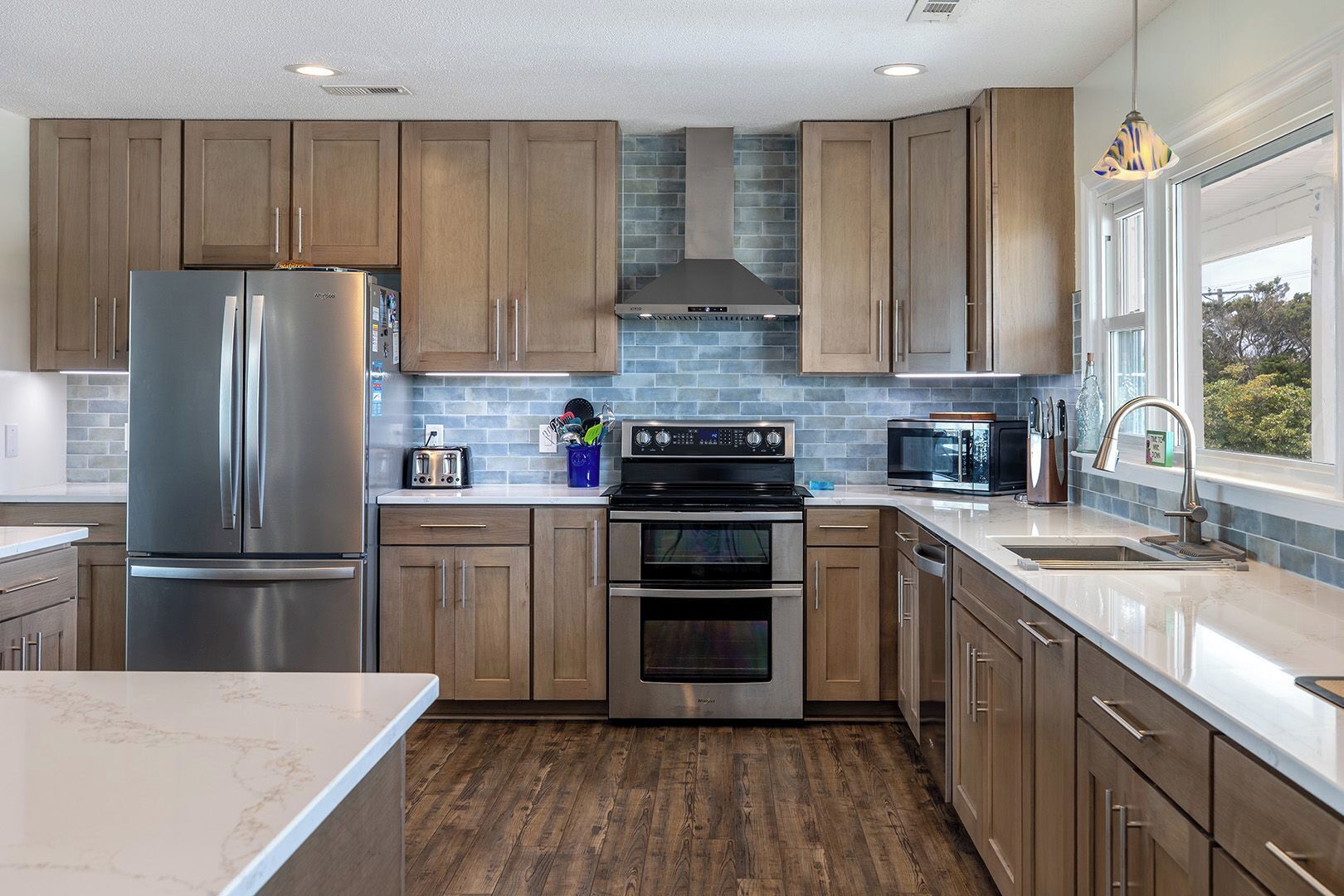 A modern kitchen with wooden cabinets, stainless steel appliances, a brick backsplash, and a large window providing natural light.