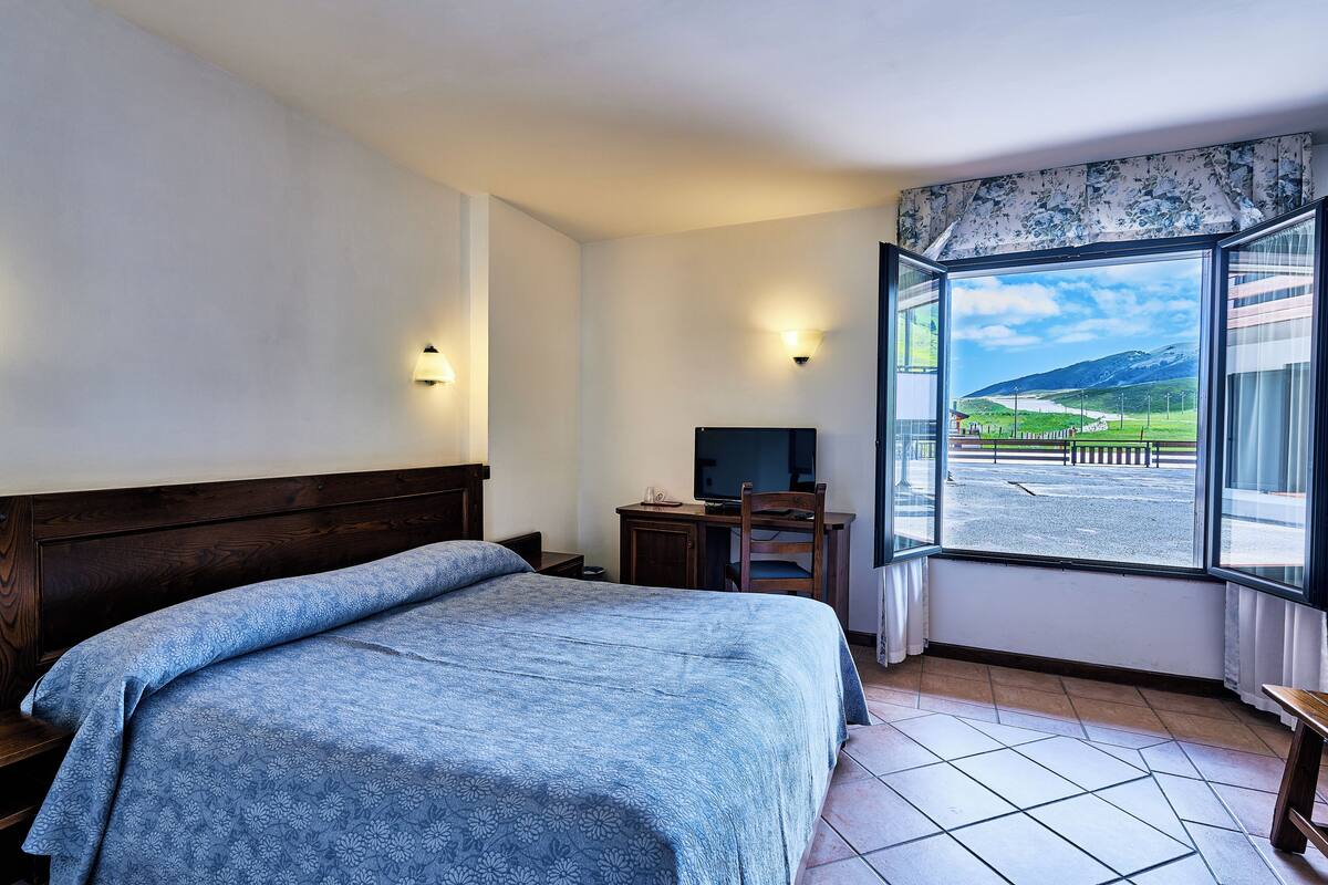 A bedroom with a large bed, a desk, a chair, and a television. There is a window with a view of a landscape.