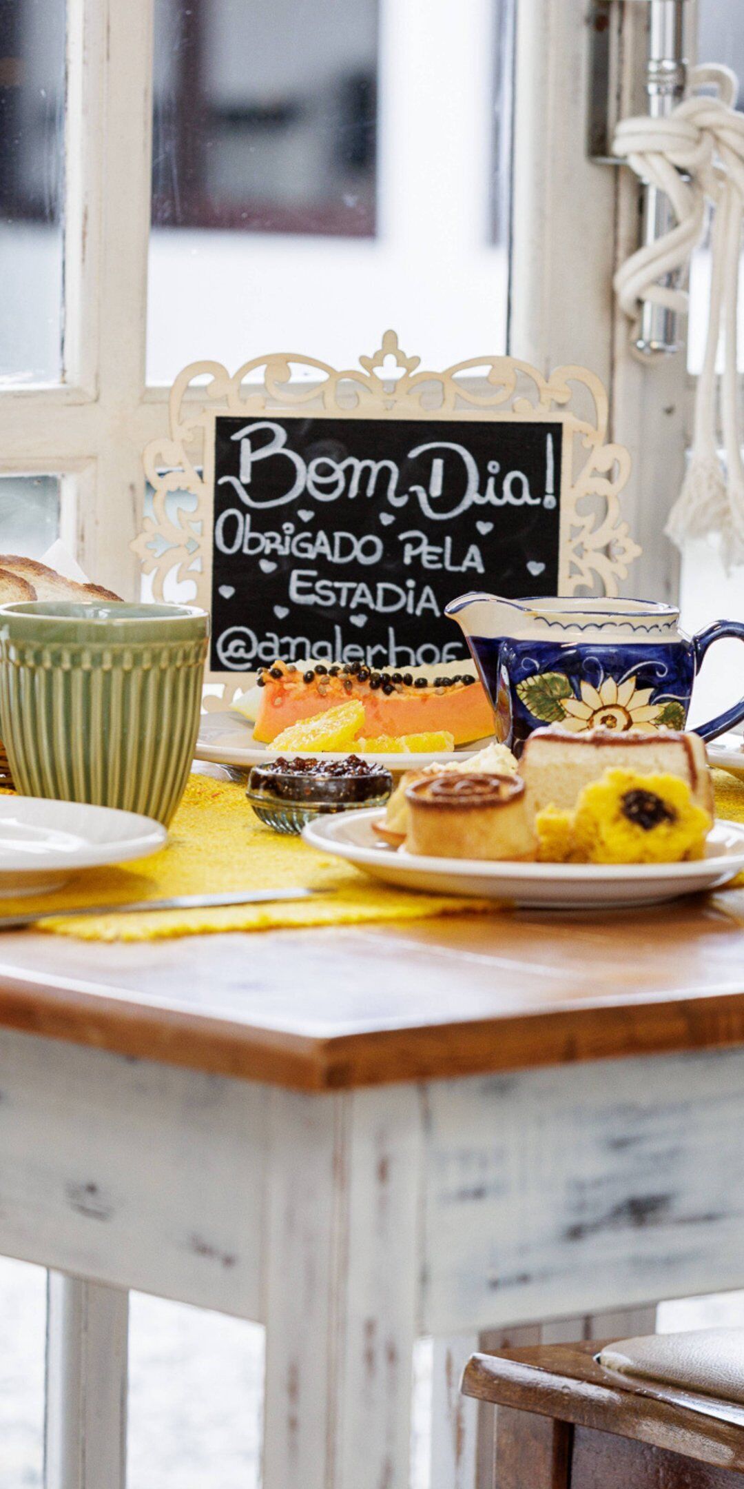 A breakfast table with various pastries and a sign that reads "Bem Dia! Obrigado pela estadia @anangelhos".