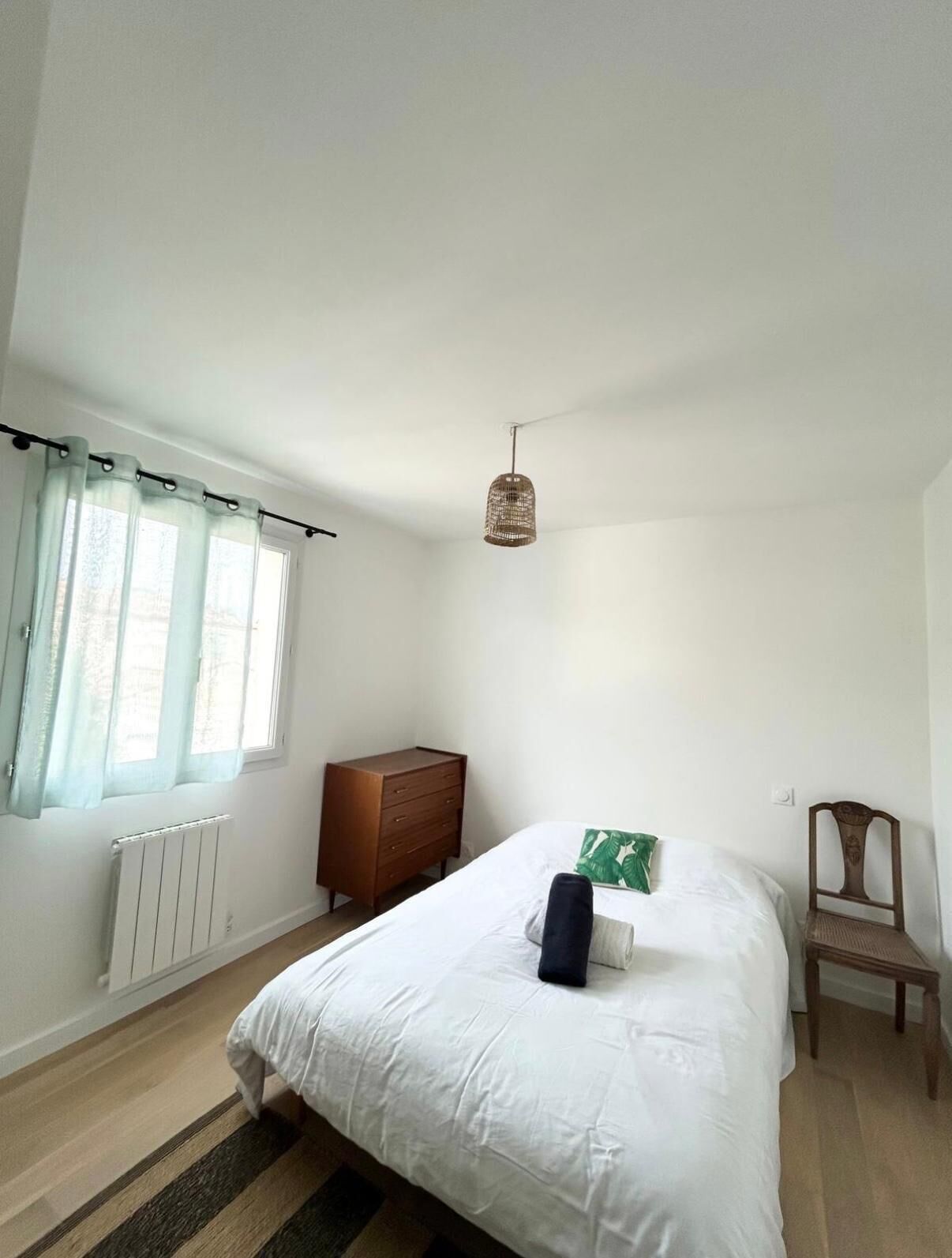 Un dormitorio con una cama, un aparador, una silla, una lámpara colgante y una ventana con cortinas.