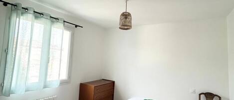 Un dormitorio con una cama, un aparador, una silla, una lámpara colgante y una ventana con cortinas.