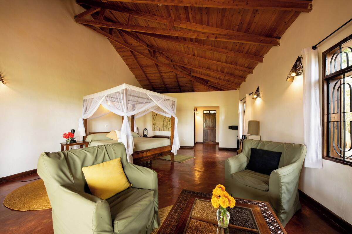 Ein Zimmer mit hoher Holzbalkendecke, einem Himmelbett, zwei grünen Sessel, einem Couchtisch mit Blumenvase und Blick durch ein Fenster mit Gitterstäben.