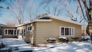 A single-story house with a gabled roof, a front porch, and a snow-covered yard. - Cozy Mid-Century Mod by Lake MI&Dunes with Hot Tub (Sawyer)