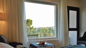 Habitació d'hotel amb una finestra gran, una cadira, una taula petita amb un plat de fruita i una làmpada.