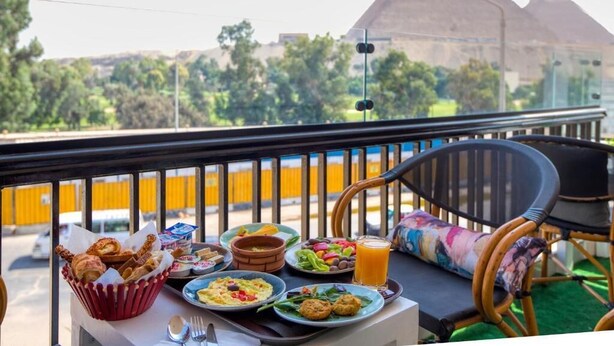 A balcony with a table set for breakfast, featuring plates of food, a basket of pastries, and a glass of orange juice. In the background, two pyramids are visible. - Maria Pyramids House - Superior King Room (Giza Governorate)