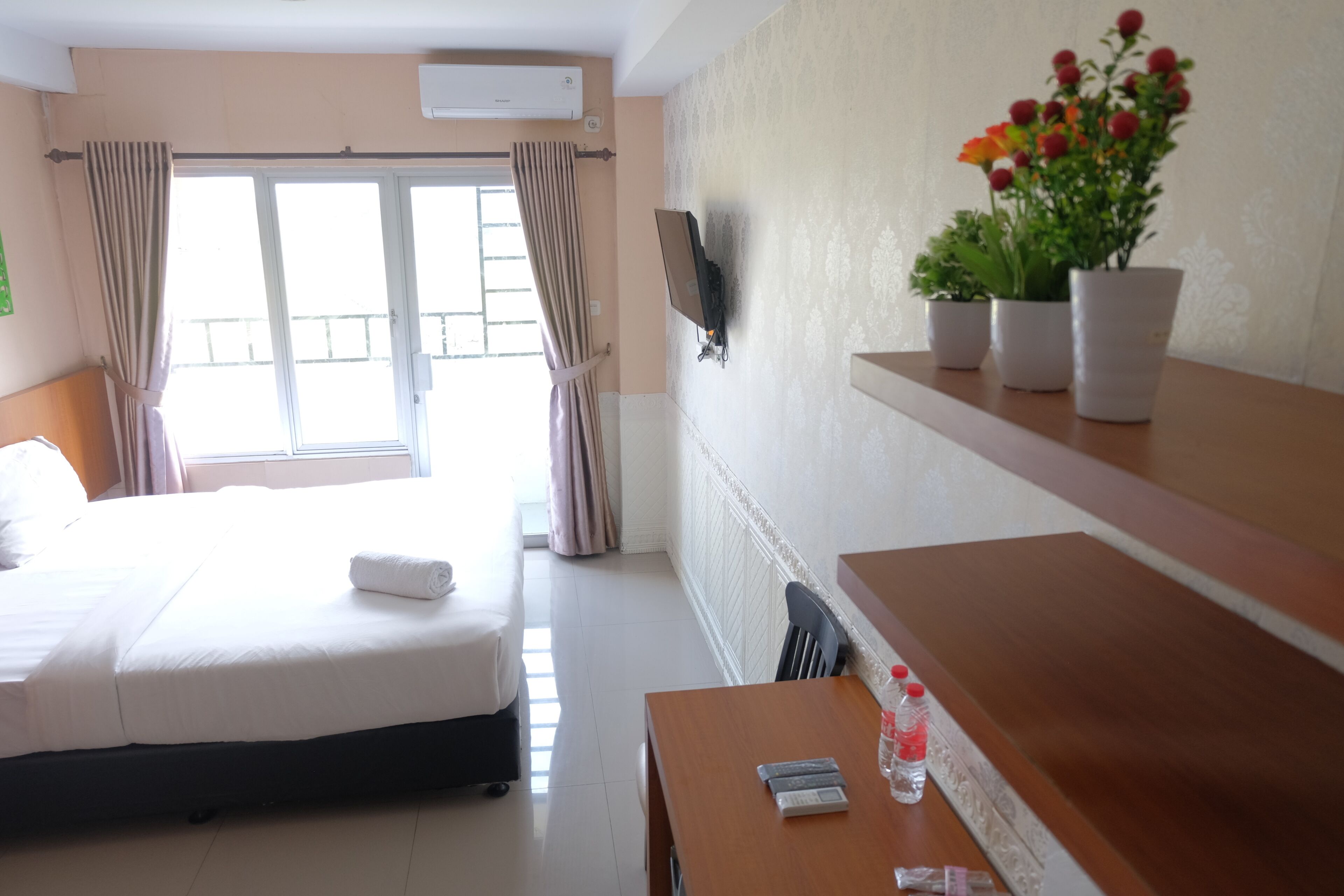 A hotel room with a bed, a table with a bottle and a remote control, a TV mounted on the wall, and a window with curtains.