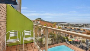 A balcony with two metal chairs, a swimming pool, and a view of a coastal town. - Hydra 8 (Merimbula)