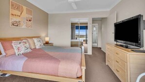 A bedroom with a bed, a TV, a ceiling fan, a nightstand, and a window with a view. - Hydra 8 (Merimbula)