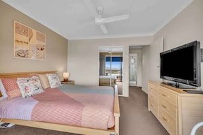 A bedroom with a bed, a TV, a ceiling fan, a nightstand, and a window with a view.