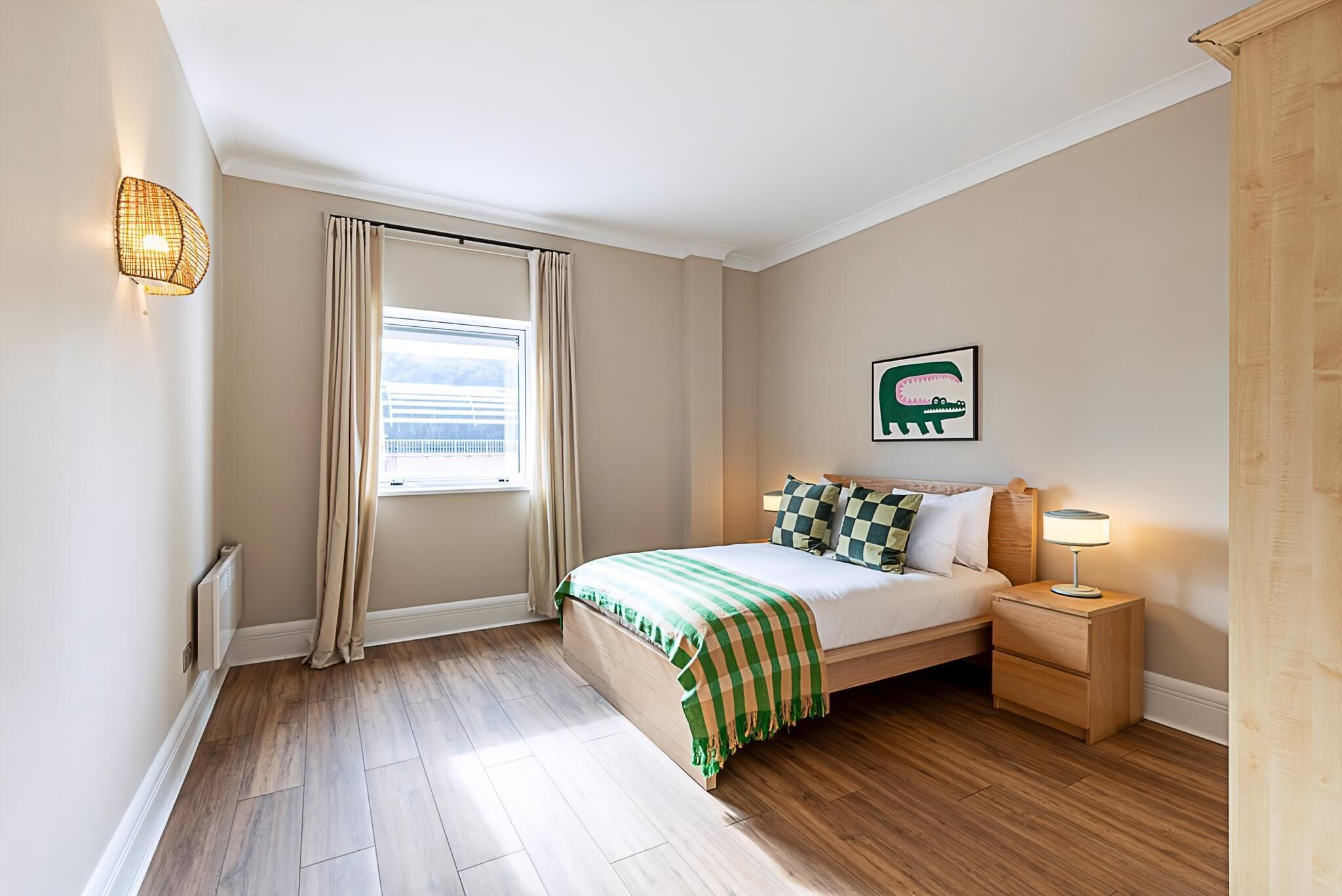A bedroom with a bed, a nightstand, a wall-mounted lamp, a window with curtains, and a picture on the wall.
