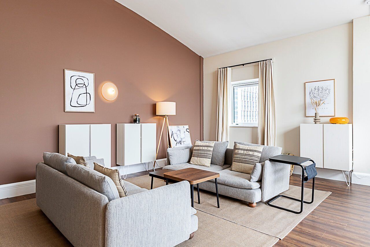 Ein modernes Wohnzimmer mit einer Sofagarnitur, einem Couchtisch und einer Lampe. An den Wänden befinden sich gerahmte Kunstwerke und ein Fenster mit Vorhängen.