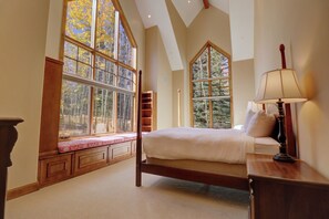 A bedroom with a large window, a bed, a bedside table with a lamp, and built-in shelving.