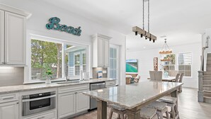 A modern kitchen with white cabinetry, a large island, and a dining area with a wooden table and chairs. - Blue Fish Bungalow (Santa Rosa Beach)