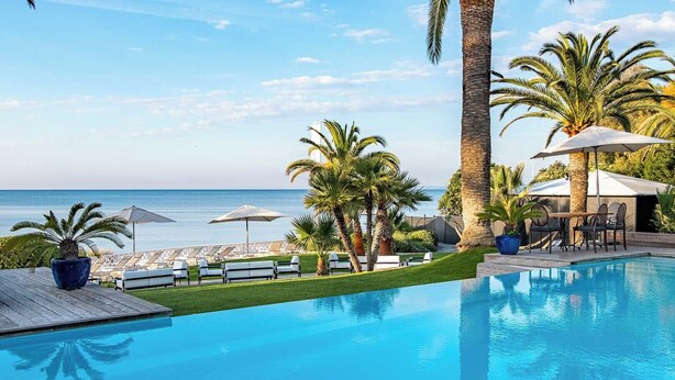 A pool area with lounge chairs and umbrellas, a view of the ocean, and palm trees.