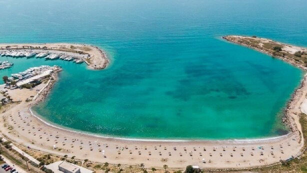 An aerial view of a coastal area with a marina, a beach with sun loungers, and a small island. - Glyfada's House Project!
 (Glifada)