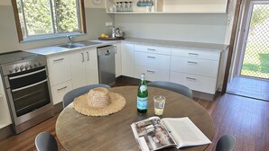 A kitchen with a stove, oven, sink, and a dining table with a magazine, a straw hat, and a bottle. - 29 Ocean Road, Brooms Head (Brooms Head)