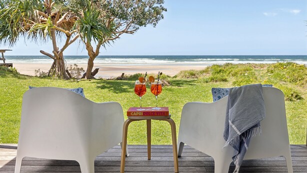 Two white chairs facing a beach with a wooden table between them, holding a book titled "CAPRI DOLCE VITA" and two glasses of red drink. - 29 Ocean Road, Brooms Head (Brooms Head)