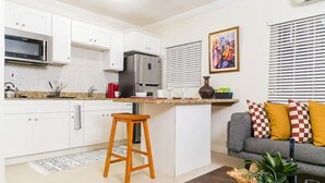 Eine moderne Küche mit weißen Schränken, einem Edelstahlkühlschrank und einer Mikrowelle. Ein Holzschemel und ein graues Sofa mit karierten Kissen sind ebenfalls vorhanden.