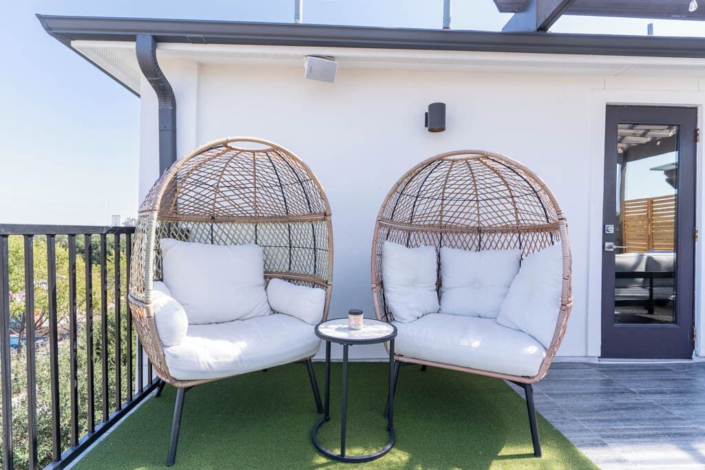 Two wicker chairs with white cushions on a balcony with artificial grass, a small round table, and a door in the background.