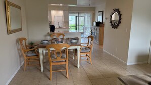 Un comedor con una mesa dispuesta para cuatro personas, rodeado de sillas de madera con asientos acolchados. Se observa una cocina con un fregadero y armarios al fondo.