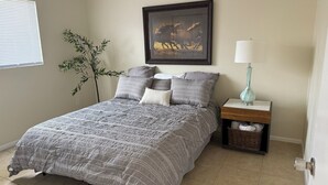 Un dormitorio con una cama, almohadas, una mesita de noche, una lámpara, un cuadro enmarcado y una planta.