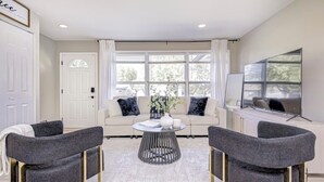 Ein modernes Wohnzimmer mit einer Couch, zwei Sesseln, einem Couchtisch und einem großen Fenster mit Vorhängen.