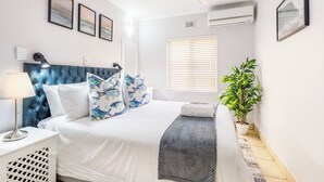 Un dormitorio con cama, mesita de noche, lámpara, ventilador de techo, ventana con persianas y una planta en maceta.