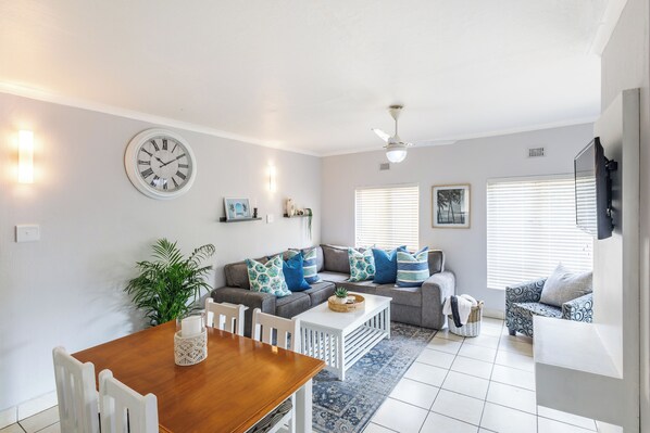 A modern living room with a wooden dining table, white chairs, a grey sofa with blue and white cushions, a blue patterned rug, and a wall clock.