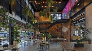A modern indoor dining area with multiple wooden tables and chairs, hanging plants, and a neon sign.