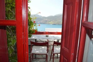 An outdoor seating area with a table and chairs, a red door, and a view of a body of water and hills. - The Red-Fish House - 1 (Astipalea)