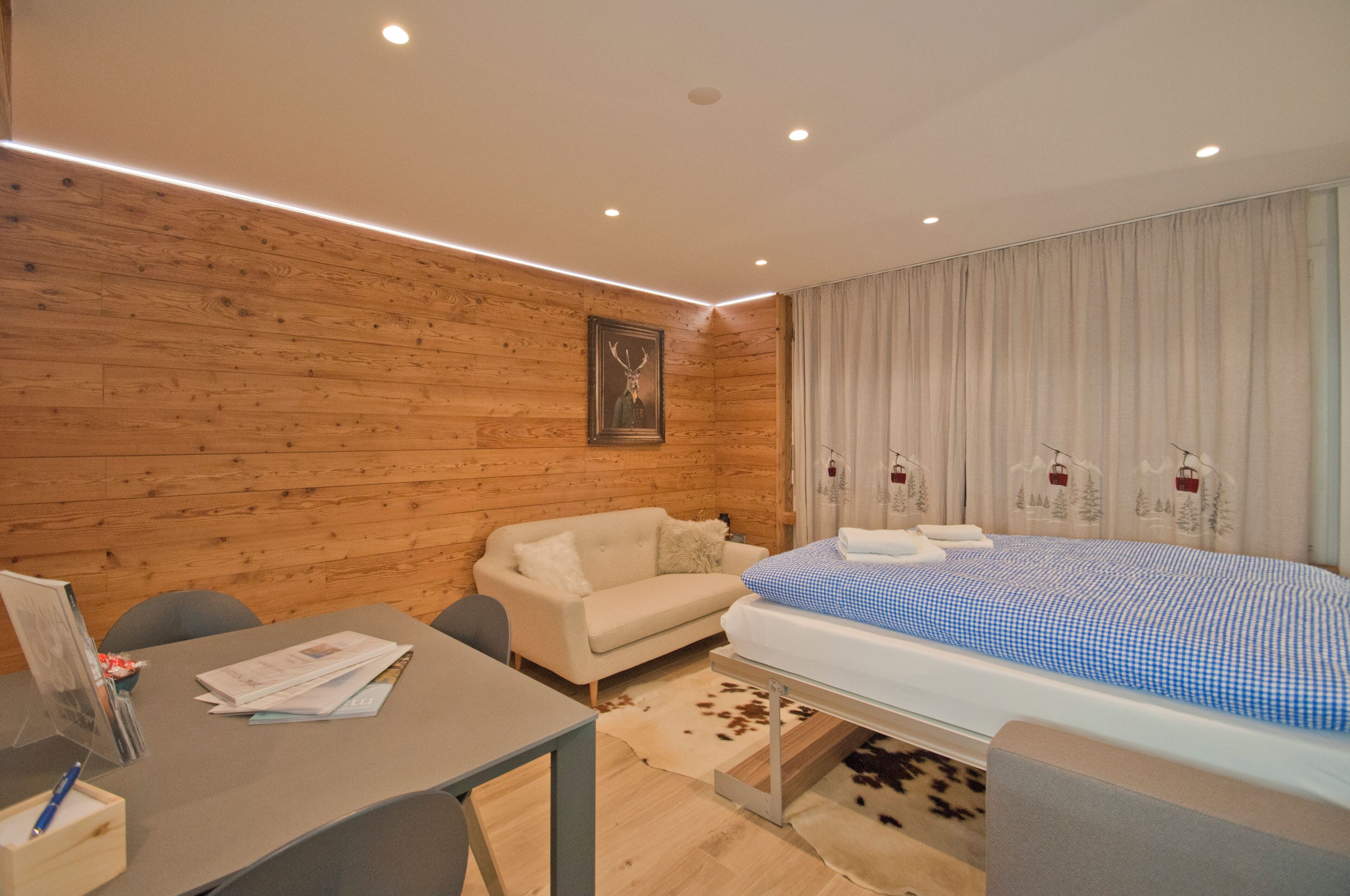 A modern hotel room with wooden panel walls, a bed with a checkered blue and white bedspread, a white sofa, a small table with a pen and papers, and a cowhide rug.