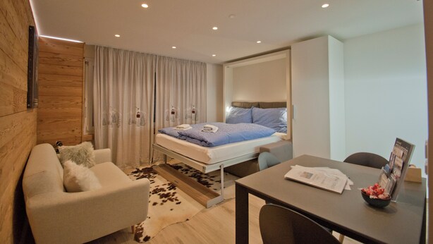 A modern hotel room with a bed, a sofa, a dining table, and a small kitchen area. - Studio Aosta (Zermatt)