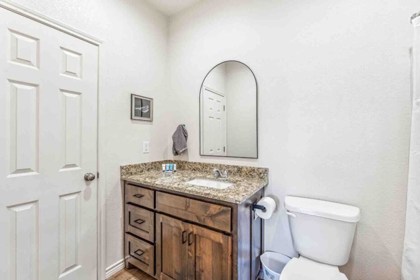 A bathroom with a white door, a granite countertop sink, wooden cabinets, a mirror, and a toilet.