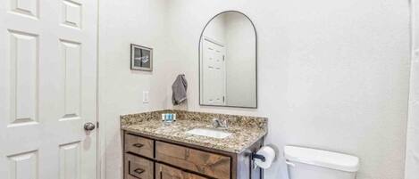 A bathroom with a white door, a granite countertop sink, wooden cabinets, a mirror, and a toilet.