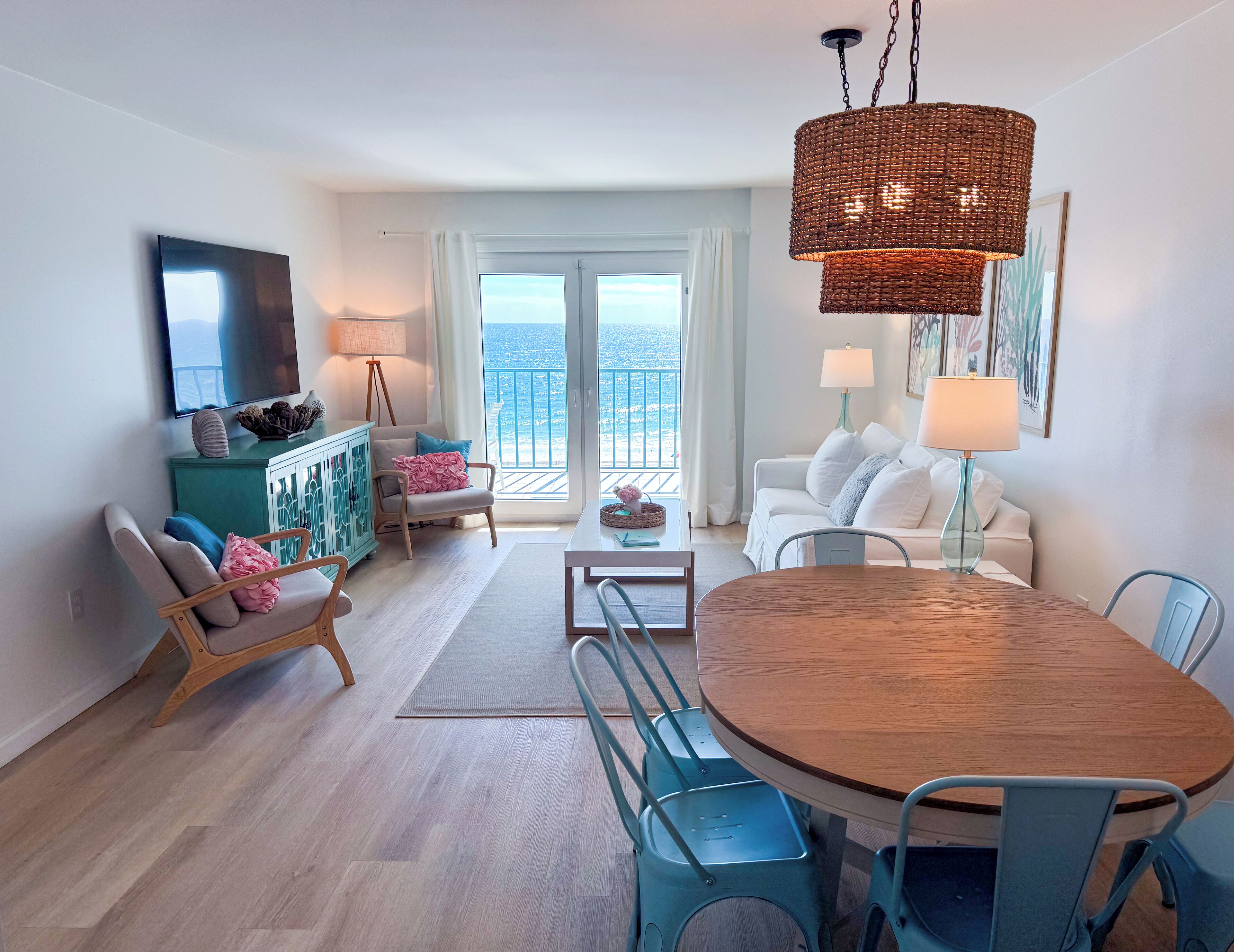 A modern living room with a wooden dining table, blue chairs, a white sofa, and a view of the ocean through large windows.