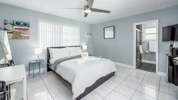 A modern hotel room with a bed, a ceiling fan, a TV, and a bathroom visible through an open door. - Cozy Getaway Studio with Pool and Putting Green 1A (Fort Lauderdale)