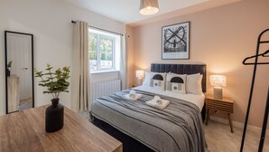A neatly arranged bedroom with a large bed, bedside lamps, a bedside table, a window with curtains, a potted plant, and a framed artwork on the wall. - Spacious 5-Bedroom House in Nottingham (Nottingham)