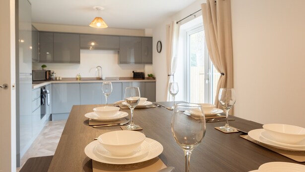A modern kitchen with grey cabinets, a wooden dining table set for four with white plates and wine glasses, and pendant lights. - Spacious 5-Bedroom House in Nottingham (Nottingham)