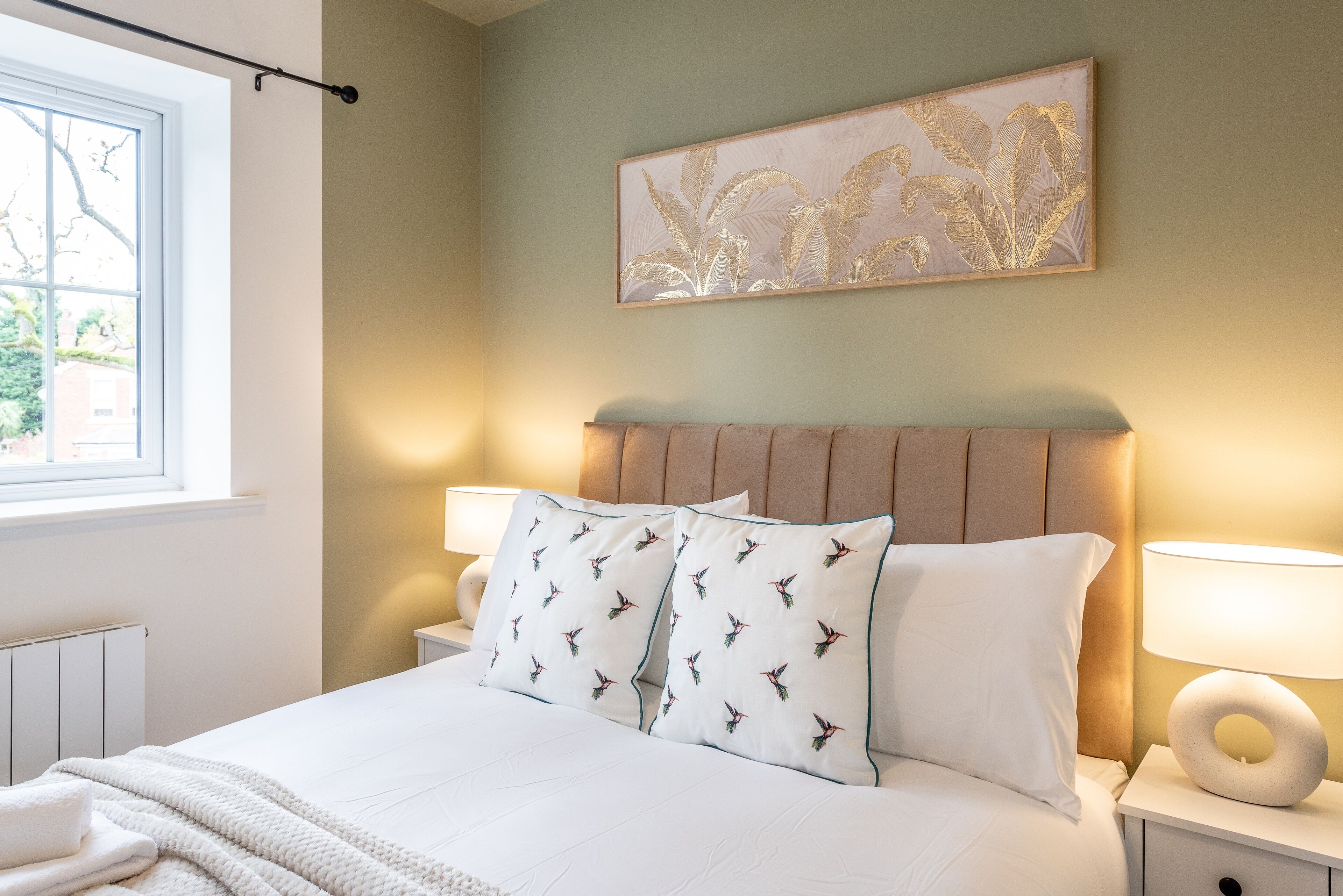 A neatly made bed with white linens and patterned pillows, a bedside lamp, and a wall-mounted artwork.