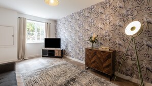 A modern living room with a flat-screen TV, a wooden cabinet, a standing lamp, and a patterned wallpaper. - Spacious 5-Bedroom House in Nottingham (Nottingham)