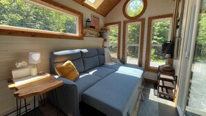 Et hyggeligt sommerhusinteriør med en sofa, et lille bord og et vindue med udsigt til en skov.