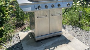 En udendørs grill i rustfrit stål med fire brændere, en tilhørende hylde og et kontrolpanel, placeret på en støbt betonplade i et skovområde.