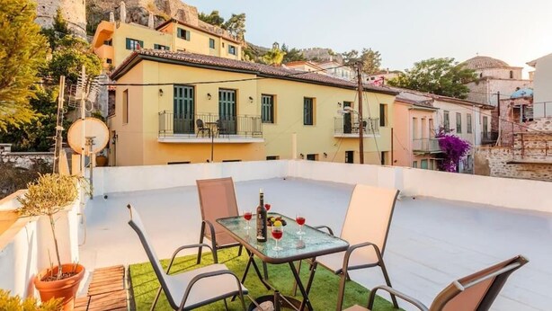 A rooftop terrace with a table set for dining, surrounded by chairs, overlooking a hillside with colorful buildings. - Nafplia Guesthouse 1 (Nafplio)