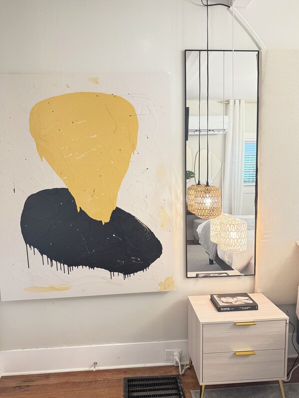 Quarto moderno com uma grande pintura abstrata, uma mesa de cabeceira com um livro e um espelho refletindo uma cama e luminárias pendentes.